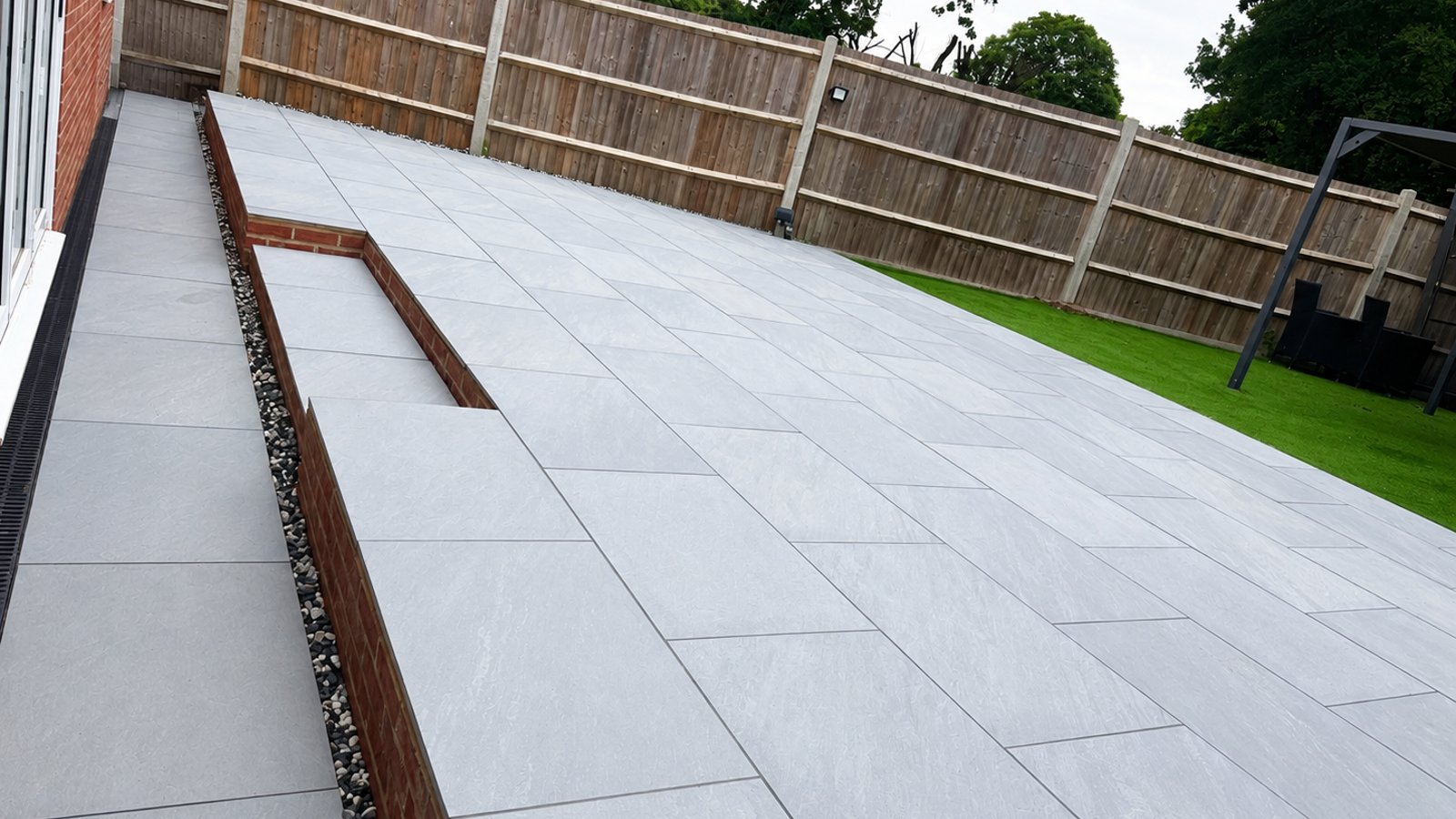 Grey porcelain patio with brick-edged pebble border and lawn.